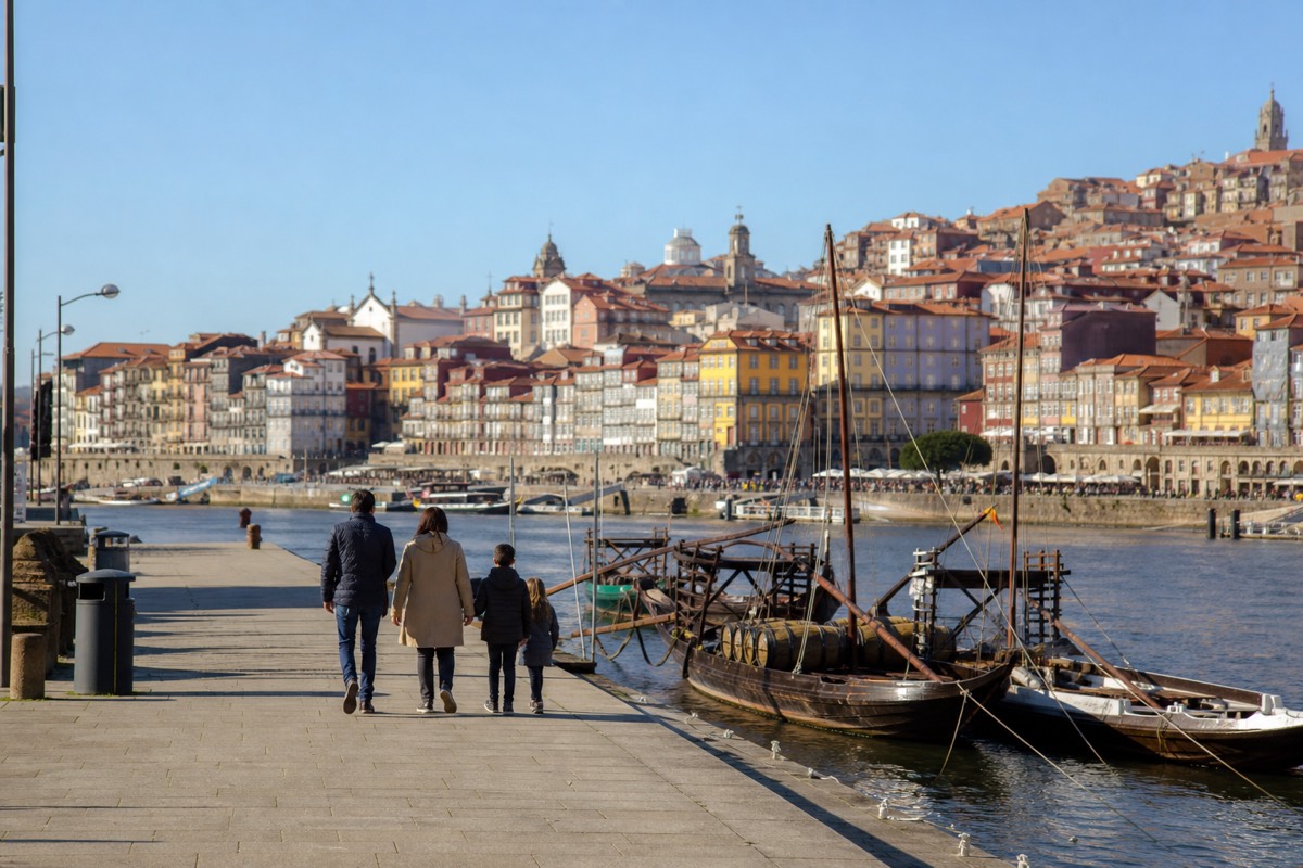 Family walking along the Vila Nova de Gaia riverfront with moored rabelo boats and the Dom Luís I Bridge behind them